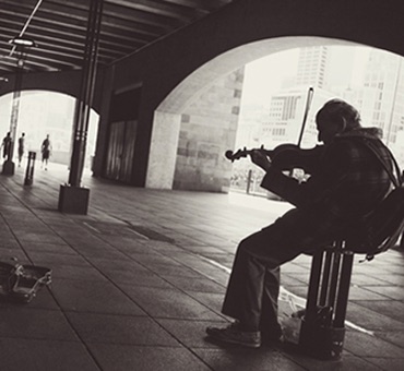 black and white photo of old man sitting down and playing violin underneath urban bridge